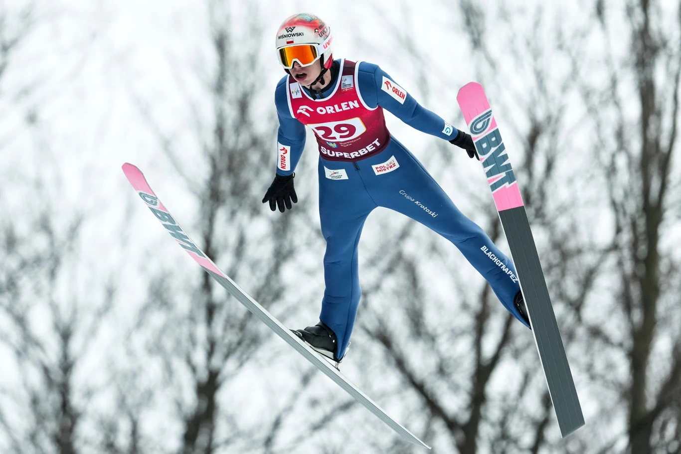Zawodnik w kombinezonie narciarskim lecący w powietrzu podczas skoku narciarskiego, w tle rozmyte sylwetki drzew, charakterystyczny sprzęt sportowy i kask z goglami.