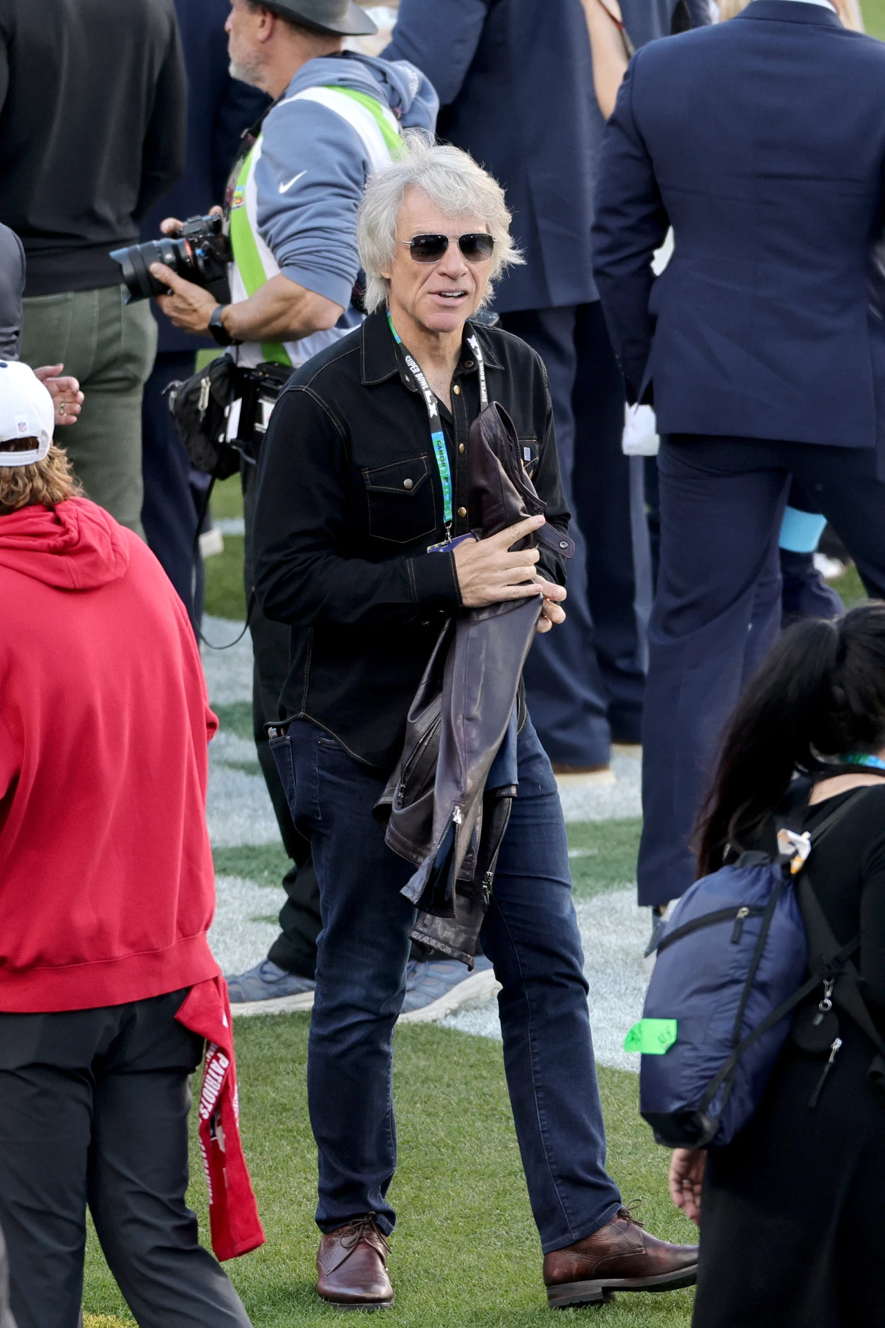 Mężczyzna w okularach przeciwsłonecznych i czarnej kurtce stoi na murawie, trzymając w ręku kurtkę. Wokół niego znajduje się kilka osób w różnokolorowych ubraniach, część z aparatem lub sprzętem medialnym.