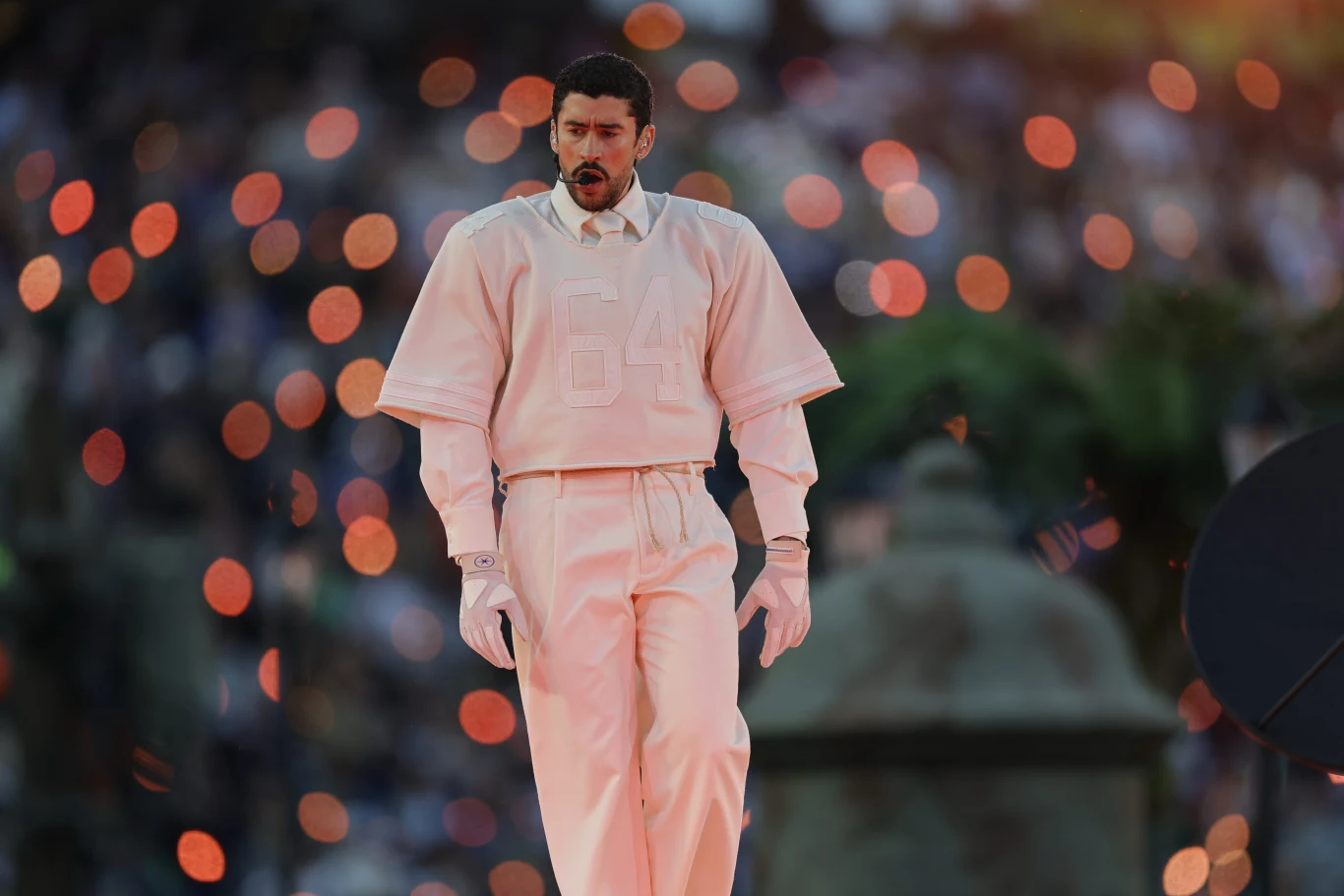 Bad Bunny wystąpił podczas Super Bowl 2026 Mężczyzna ubrany w jasny, luźny strój z numerem 64 na koszulce, wyposażony w mikrofon na głowie, stoi na tle rozmytego, kolorowego oświetlenia i rozmytej publiczności.