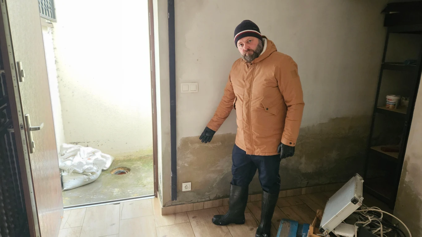 Mężczyzna w kurtce i czapce wskazuje na ścianę z widocznym śladem po zalaniu. Podłoga i część ściany są zabrudzone wodą, a na podłodze leżą zniszczone sprzęty. Otwarty drzwi prowadzi na podwórko zalane błotem.