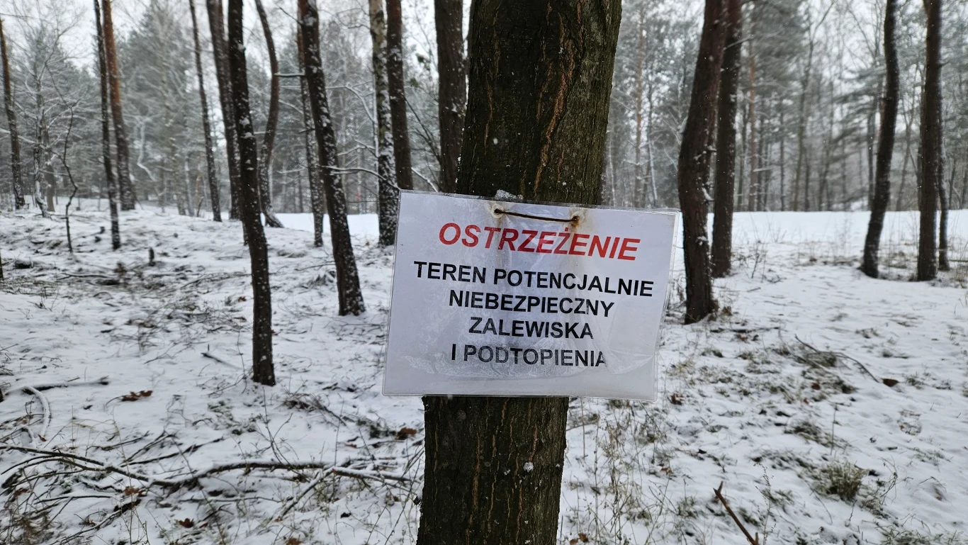 Las zimą pokryty śniegiem, na drzewie zawieszona kartka z ostrzeżeniem informującym o potencjalnie niebezpiecznym terenie, zalewisku i możliwych podtopieniach.