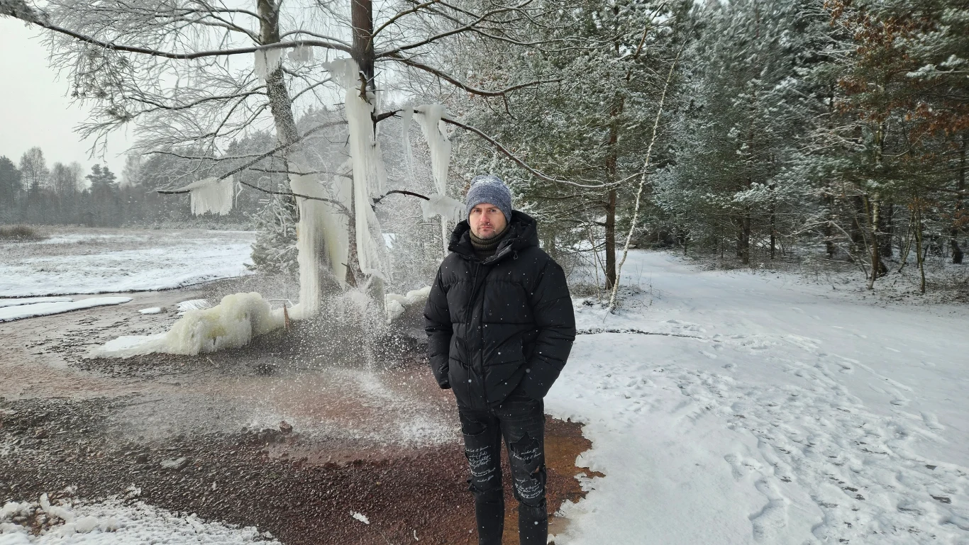 Mężczyzna ubrany w zimową kurtkę, czapkę i rękawiczki stoi na tle pokrytych śniegiem drzew oraz zamarzniętego strumienia z lodowymi formacjami na gałęziach. W tle widoczna zimowa, leśna sceneria.