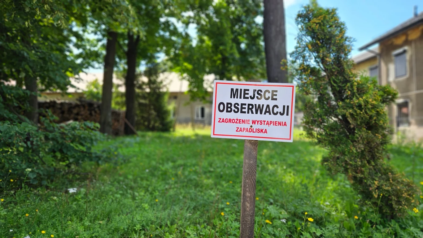 Tabliczka z napisem 'Miejsce obserwacji, zagrożenie wystąpienia zapadliska' umieszczona na tle zielonego terenu z drzewami i budynkiem w tle, ostrzegająca przed możliwym zagrożeniem.