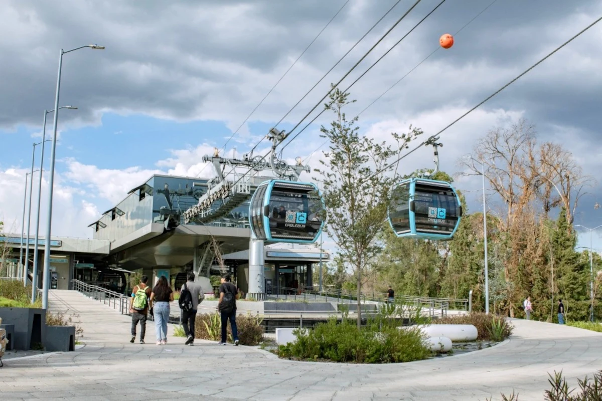 Najdłuższa miejska kolej linowa. Cablebús Línea 5 powstanie w Meksyku. Stacja kolejki linowej z unoszącymi się wagonikami, kilka osób idzie pieszo w stronę wejścia, nowoczesna infrastruktura otoczona zielenią, pochmurne niebo w tle.