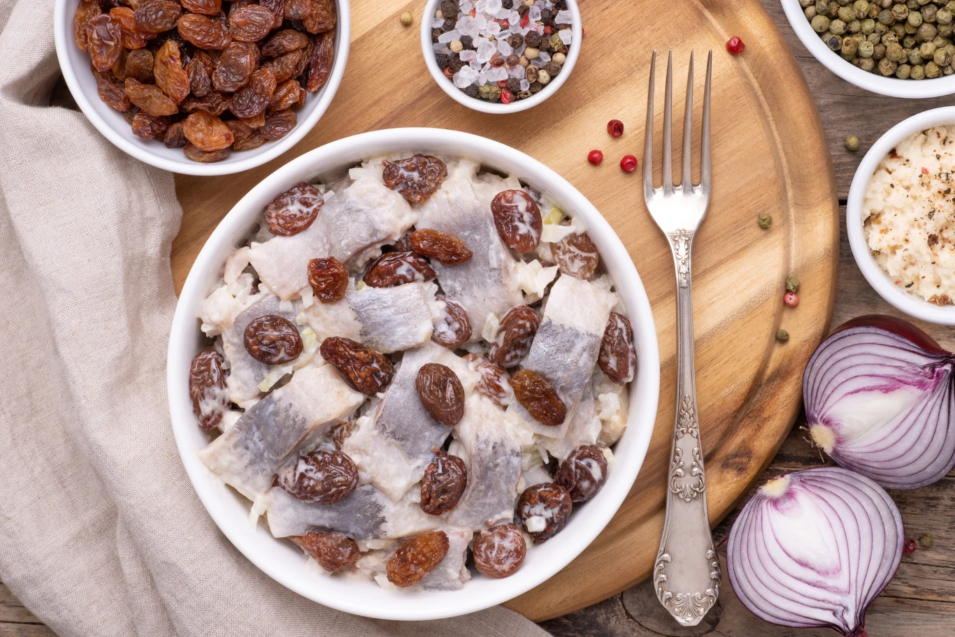 Porcja śledzi z rodzynkami ułożona w białej misce, obok plastry czerwonej cebuli, pieprz kolorowy w młynku, miseczka z rodzynkami i przyprawy na drewnianej desce, całość otoczona lnianą serwetką i widelcem.