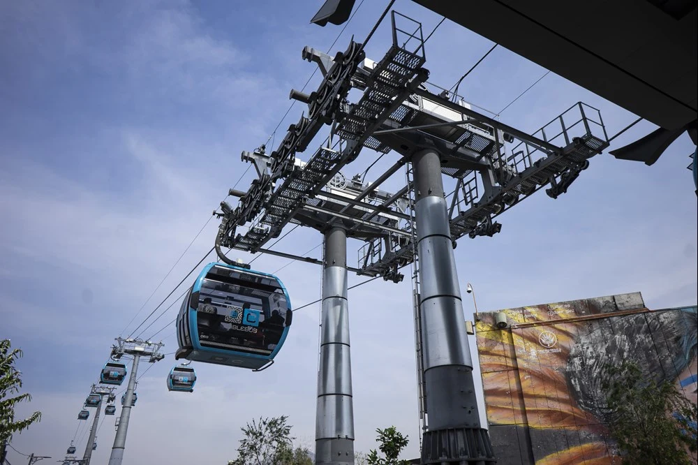 Cablebús Línea 5 będzie najdłuższą miejską koleją linową na świecie. Nowoczesna kolejka linowa zawieszona na wysokich podporach transportuje pasażerów w przeszklonych wagonikach, a w tle widoczne są elementy miejskiej zabudowy oraz fragment muralu na ścianie budynku.
