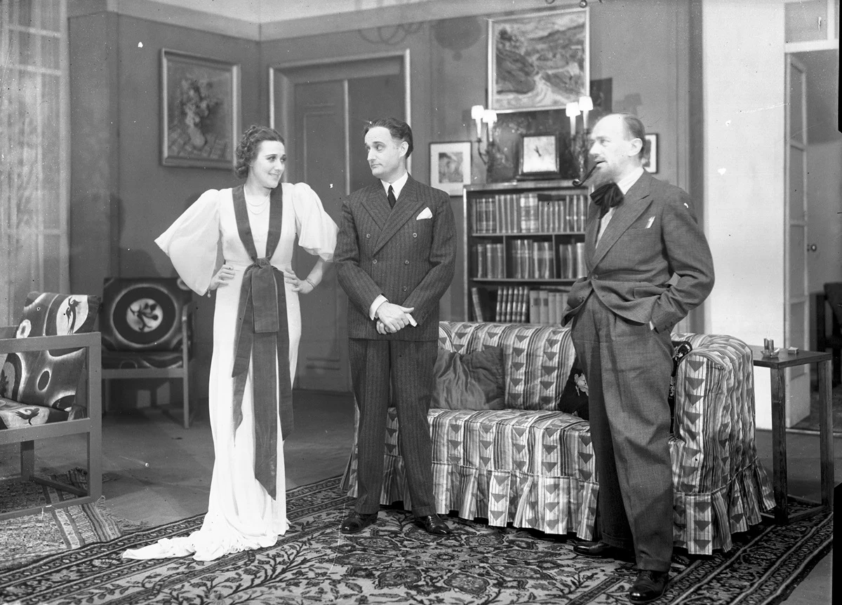 Trzy elegancko ubrane osoby rozmawiają w luksusowym salonie z bogato zdobionymi meblami, obrazami na ścianach i regałami pełnymi książek; kobieta stoi z rękami na biodrach, mężczyźni uśmiechają się.