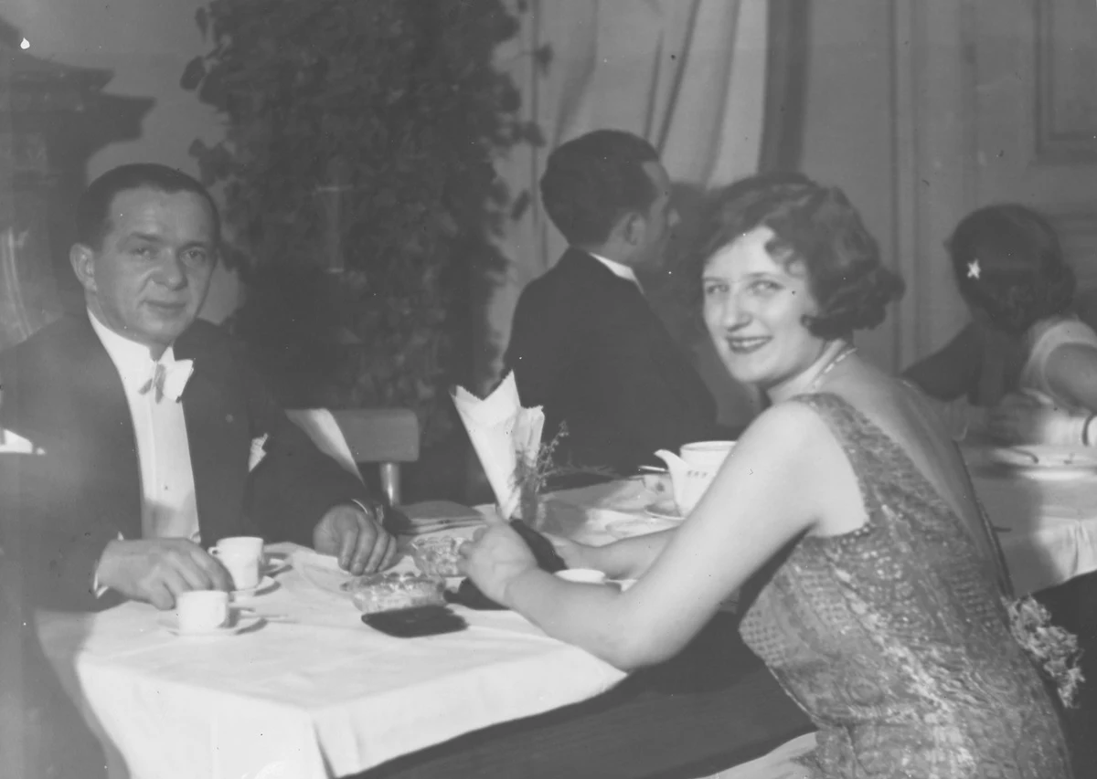 Para ubrana w eleganckie stroje siedzi przy stole w restauracji, rozmawiając i popijając kawę, w tle widoczne inne osoby oraz elegancki wystrój wnętrza.