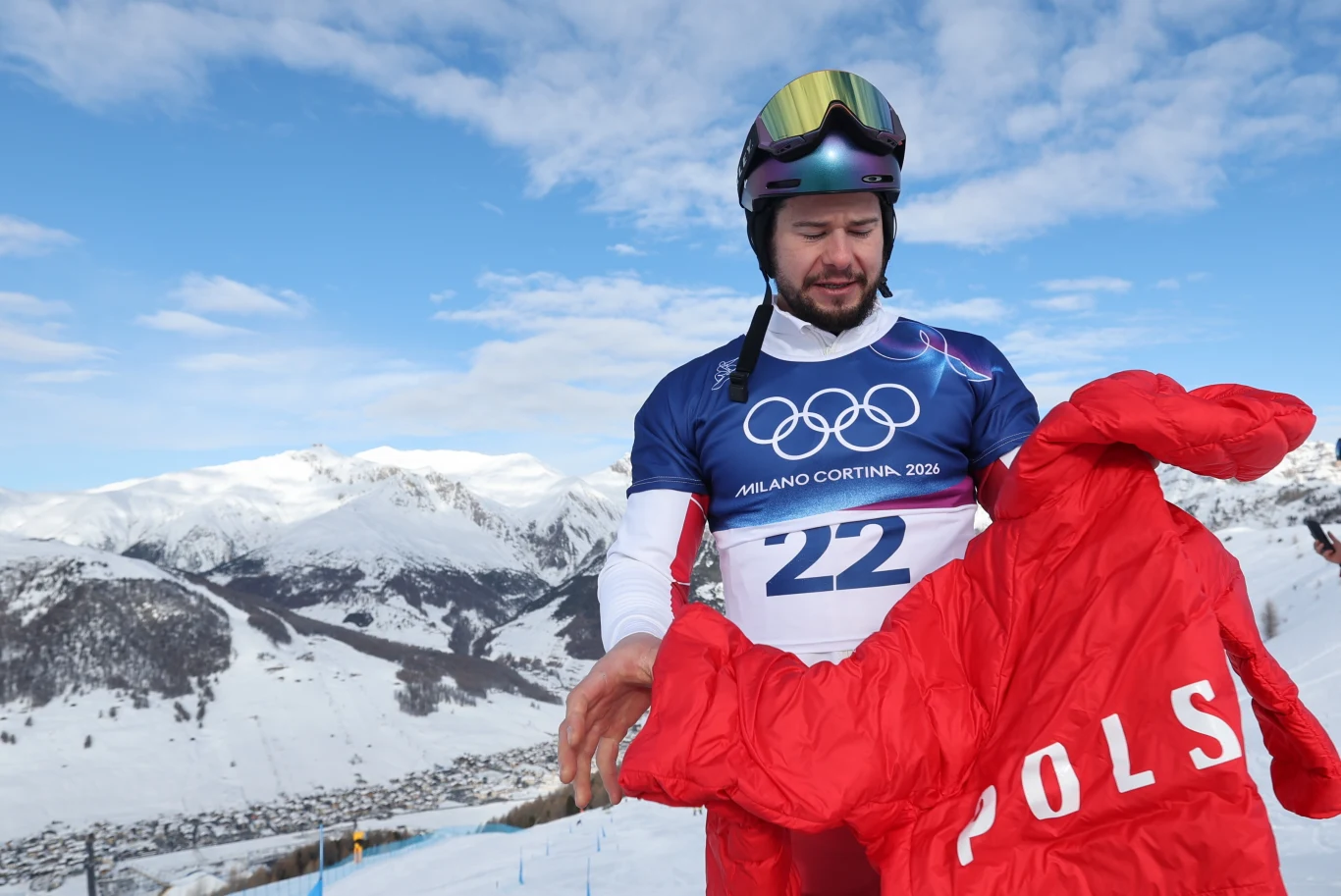 Oskar Kwiatkowski Narciarz w stroju olimpijskim na tle zaśnieżonych gór, w dłoni trzyma czerwony kombinezon z napisem 'Polska', wokół zimowy krajobraz i błękitne niebo.
