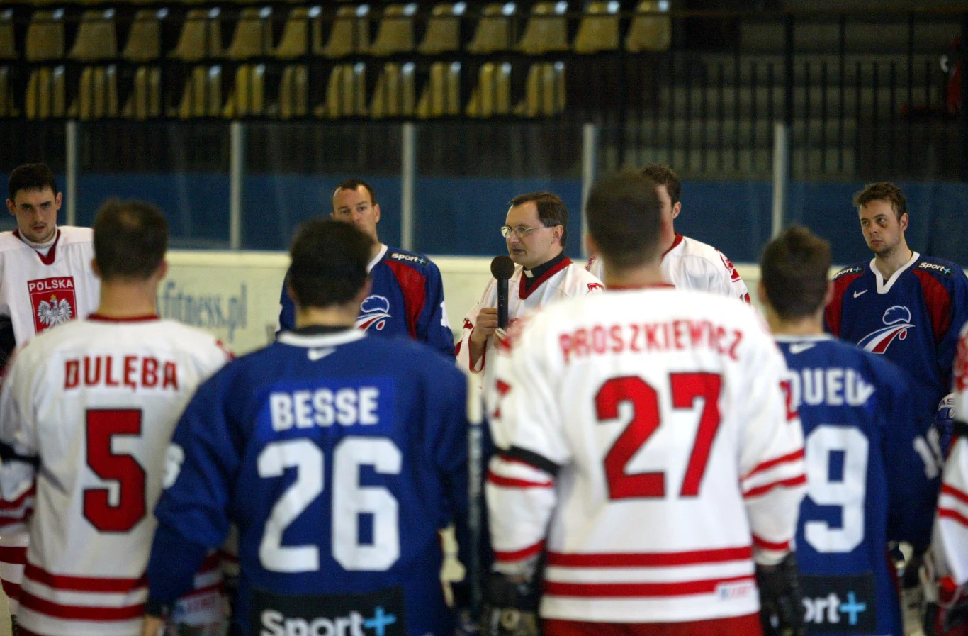 Ks. Paweł Łukaszka podczas meczu Polska - Francja (2005) Hokeiści w białych i niebieskich strojach zgromadzeni wokół trenera na lodowisku, widoczne nazwiska na plecach koszulek zawodników, w tle puste trybuny.