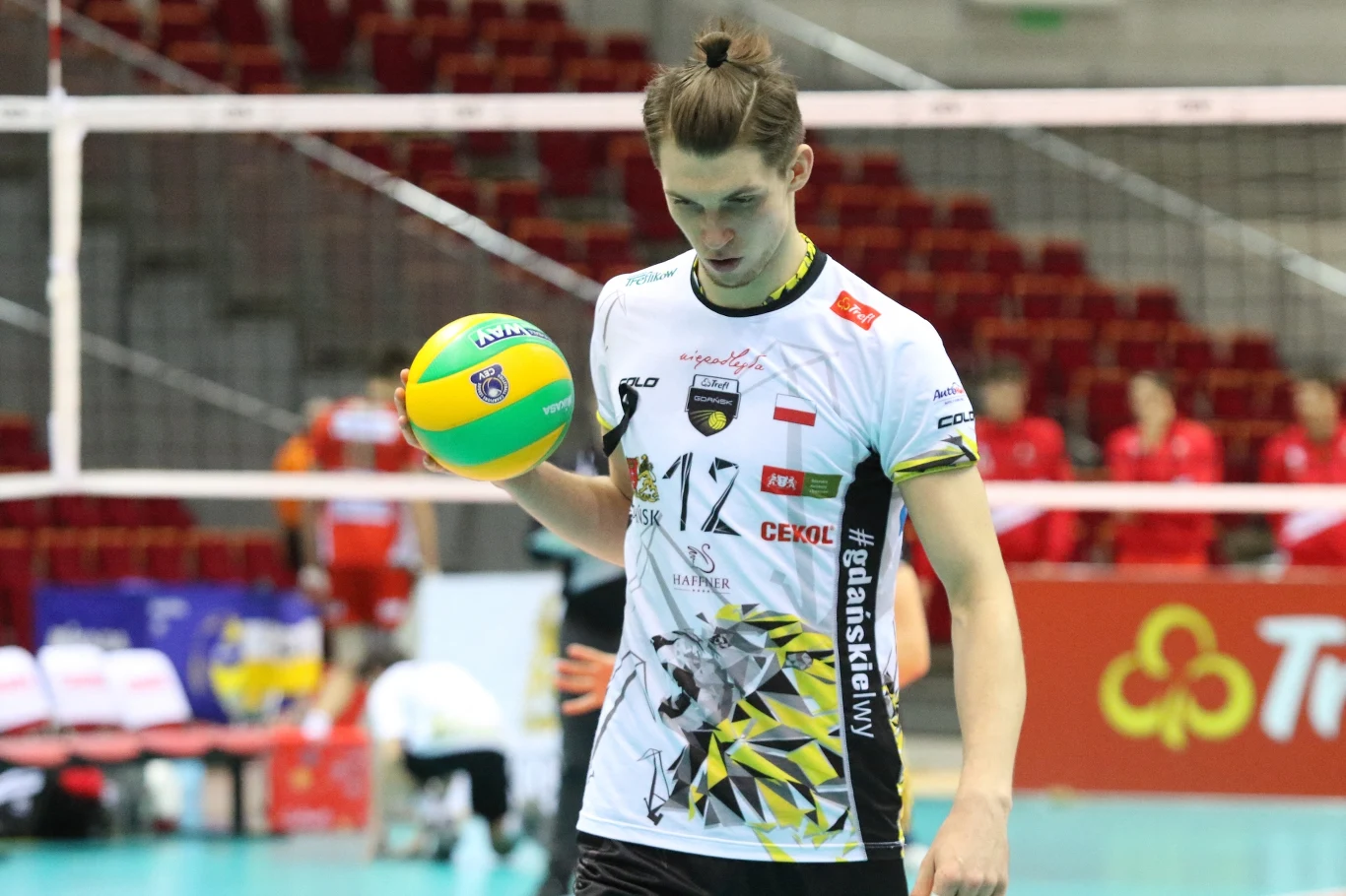 Siatkarz w sportowym stroju drużynowym z piłką do siatkówki w dłoni stoi na hali sportowej. W tle widoczna siatka oraz rozmazani członkowie drużyny po przeciwnej stronie boiska.