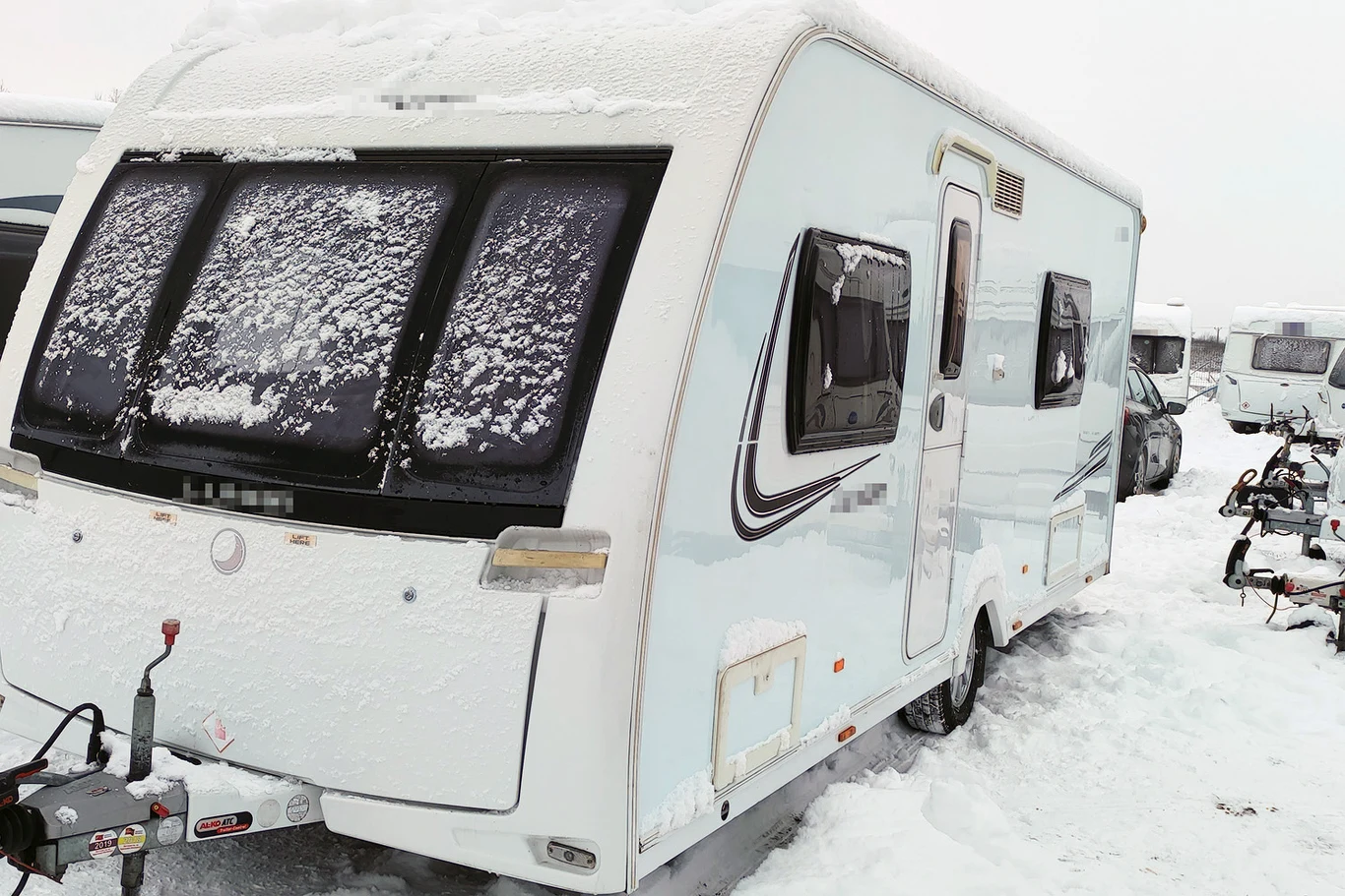 Przyczepa kempingowa pokryta śniegiem stoi na zaśnieżonym placu wśród innych podobnych pojazdów. Szyby przyczepy są częściowo zasłonięte warstwą śniegu, a teren wokół niej jest biały od opadów zimowych.