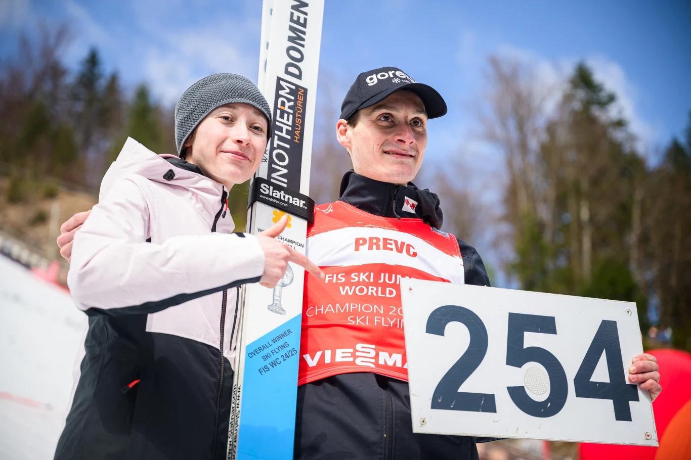 Nika i Domen Prevcowie - obecni (uznawani przez FIS) oficjalni rekordziści pod względem długości skoków Dwóch uśmiechniętych ludzi na tle zimowego krajobrazu, jedna osoba ubrana w sportowy strój z numerem 254 oraz napisem PREVC, druga w jasnej kurtce i czapce, trzymający narty do skoków narciarskich.