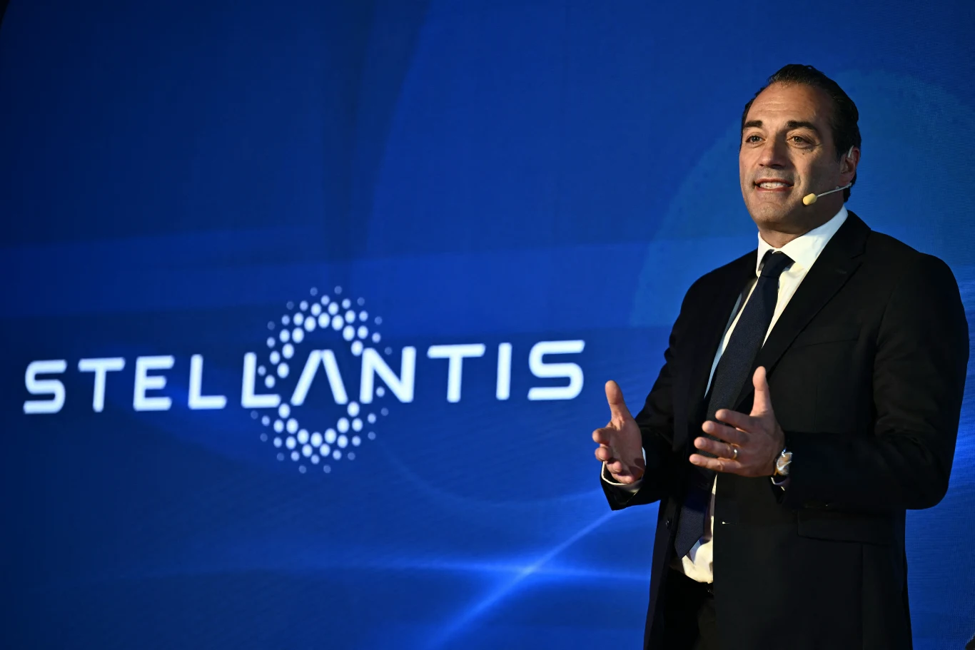 Prezes Stellantisa Antonio FIlosa zmienia kurs koncernu Mężczyzna w garniturze z mikrofonem na uchu, gestykulujący podczas prezentacji na tle niebieskiego ekranu z logo firmy Stellantis.