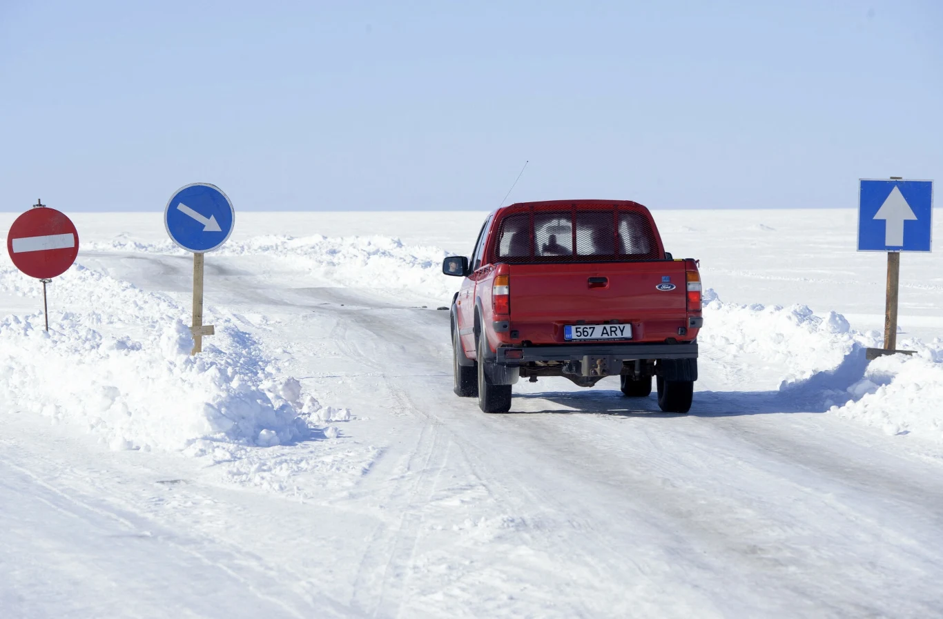 Czerwony pickup jedzie zaśnieżoną, zimową drogą pomiędzy znakami drogowymi wskazującymi zakaz wjazdu z jednej strony, nakaz jazdy w prawo oraz kierunek na wprost. Wokół droga otoczona jest grubą warstwą śniegu, niebo bezchmurne.
