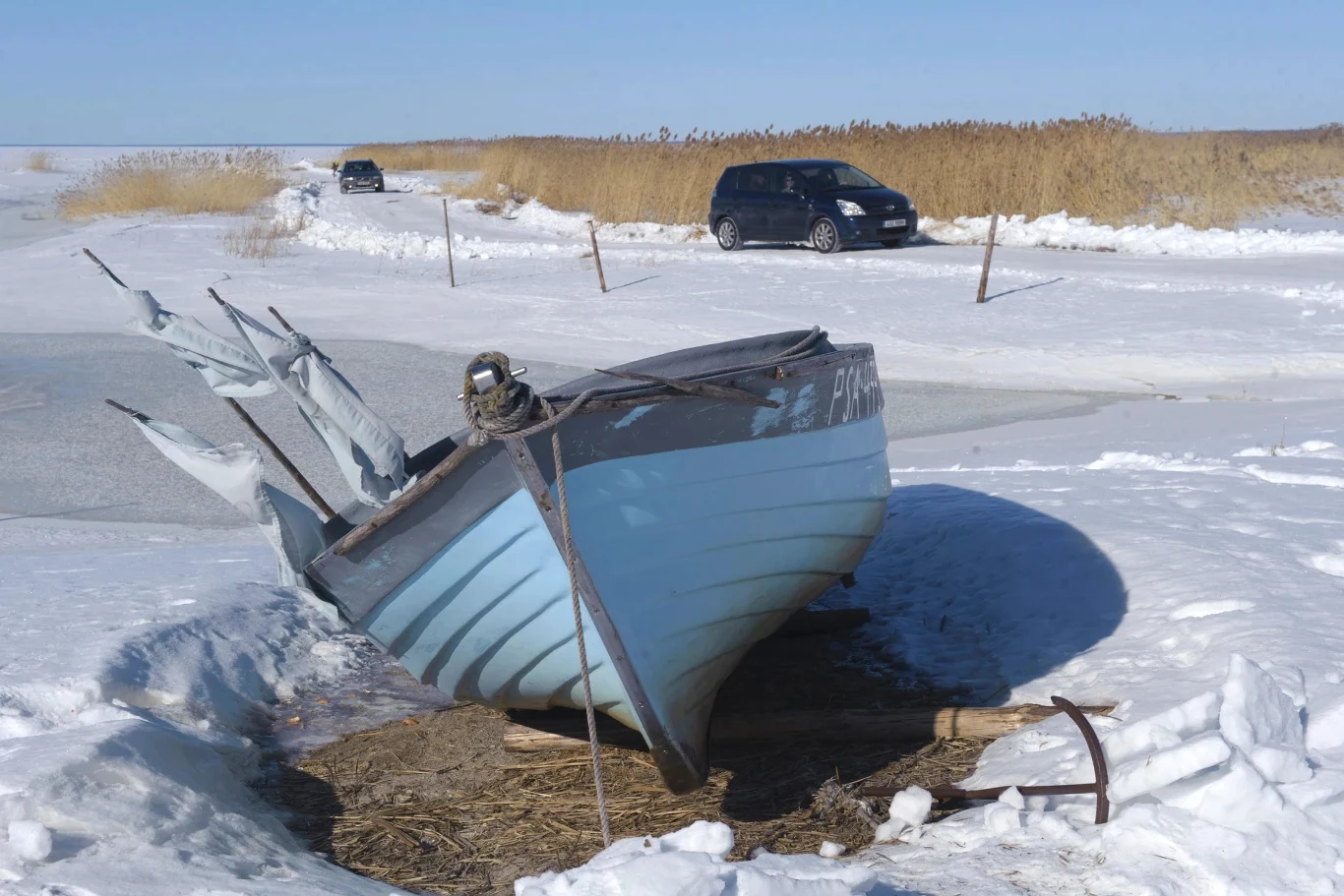 Niebieska łódź rybacka z przywiązanymi do niej sieciami stoi na zamarzniętym brzegu pokrytym śniegiem, w tle dwa samochody zaparkowane obok drogi biegnącej przez krajobraz trzcin i śniegu.