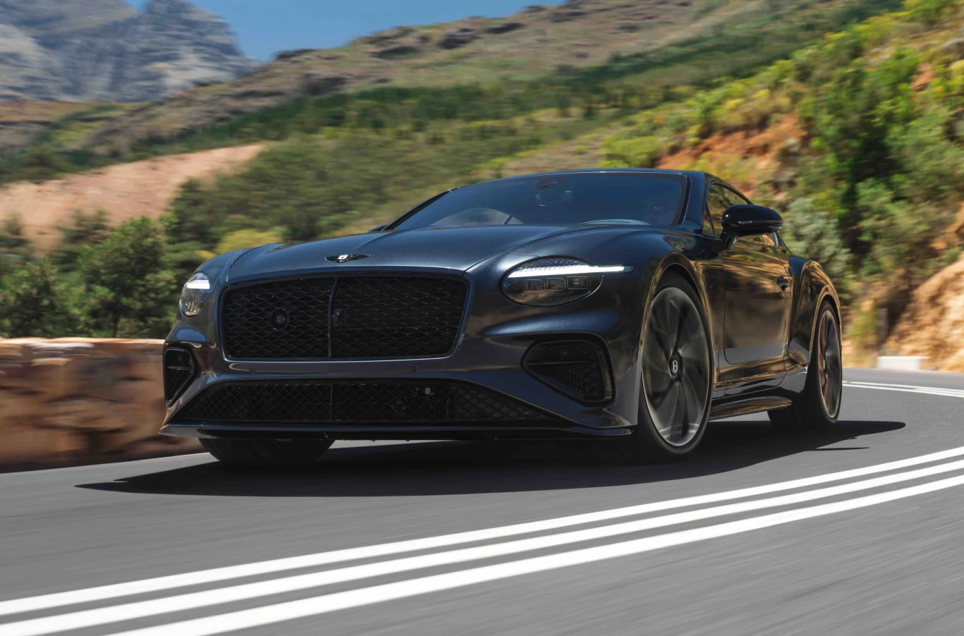Nowy Bentley Continental GT S i GTC S. 680 KM, hybryda i 306 km/h. Czarny sportowy samochód luksusowy na górskiej, krętej drodze w słoneczny dzień, w tle zielone drzewa i skaliste wzgórza.