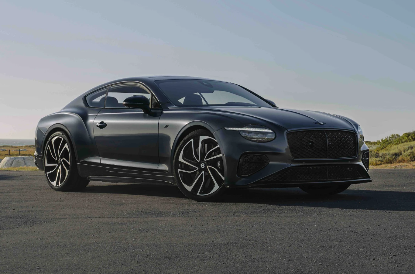 Bentley Motors wprowadził do oferty nowe wersje Continental GT S oraz GTC S. Luksusowy czarny samochód sportowy typu coupe z charakterystycznymi aluminiowymi felgami, stojący na pustym asfalcie przy naturalnym, słonecznym krajobrazie.