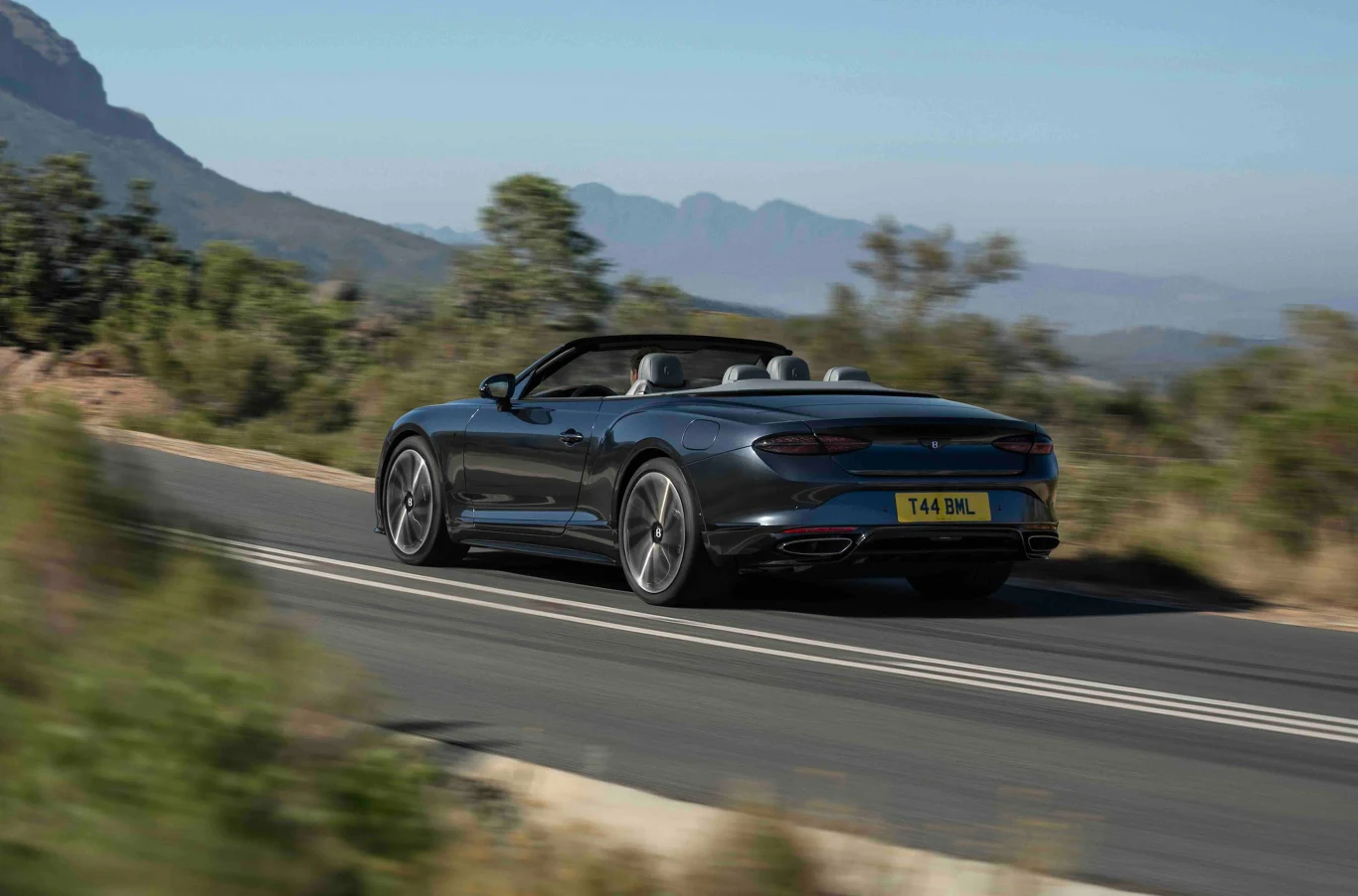 W trybie Dynamic dopuszcza kontrolowany uślizg tylnej osi, a po wyłączeniu ESC kierowca może regulować balans auta pedałem gazu. Luksusowy kabriolet jedzie asfaltową drogą przez górzysty krajobraz, otoczony przez zielone krzewy i drzewa, na tle delikatnie zamglonego pasma górskiego.