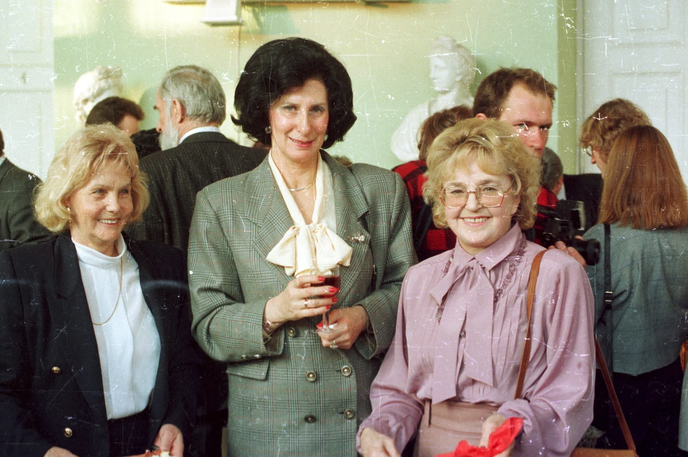 Trzy elegancko ubrane kobiety stoją razem na tle grupy ludzi zgromadzonych w jasnym pomieszczeniu, z uśmiechami na twarzach i lampką czerwonego napoju w ręku jednej z pań.