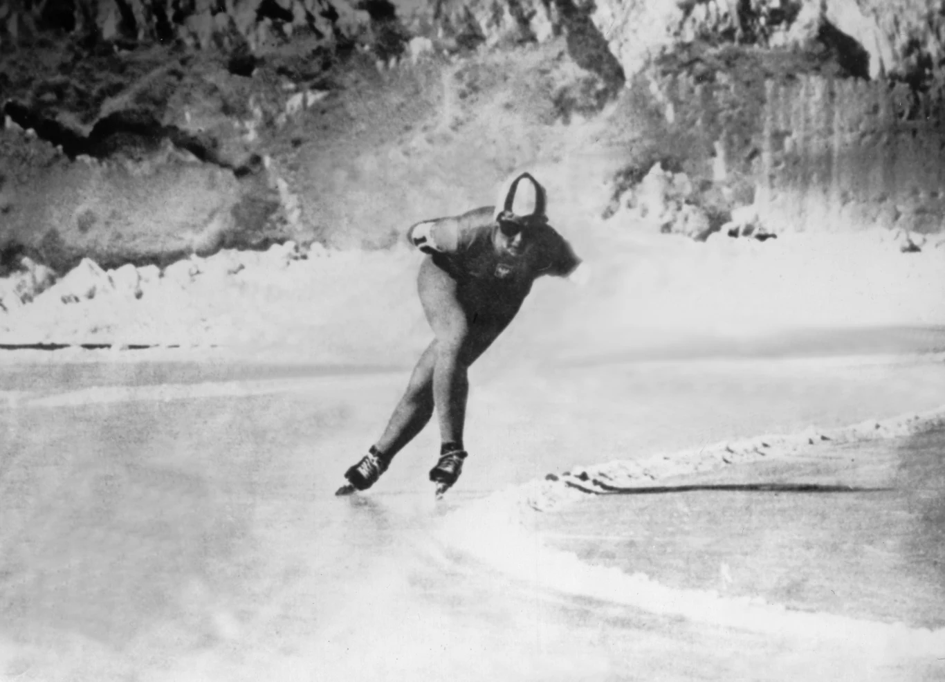 Łyżwiarz szybki w profesjonalnym stroju podczas wyścigu na zamarzniętym torze, w tle ośnieżone skały i górskie krajobrazy.