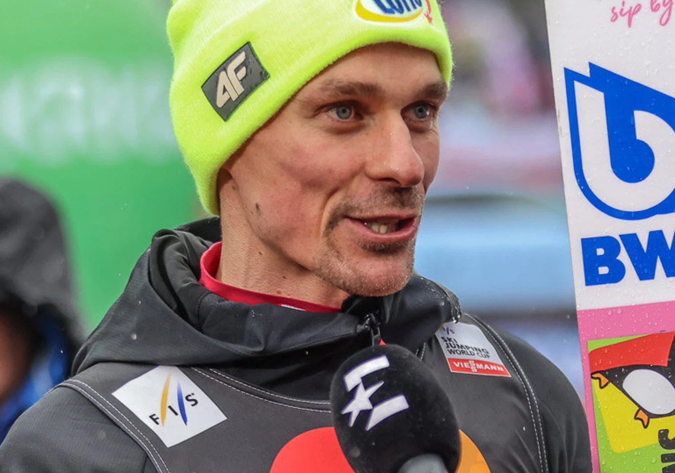 Piotr Żyła Sportowiec w czapce z logo sponsora oraz kurtce z przypiętymi identyfikatorami udziela wywiadu, trzymając deskę do skoków narciarskich oraz stojąc przed mikrofonem telewizji sportowej.