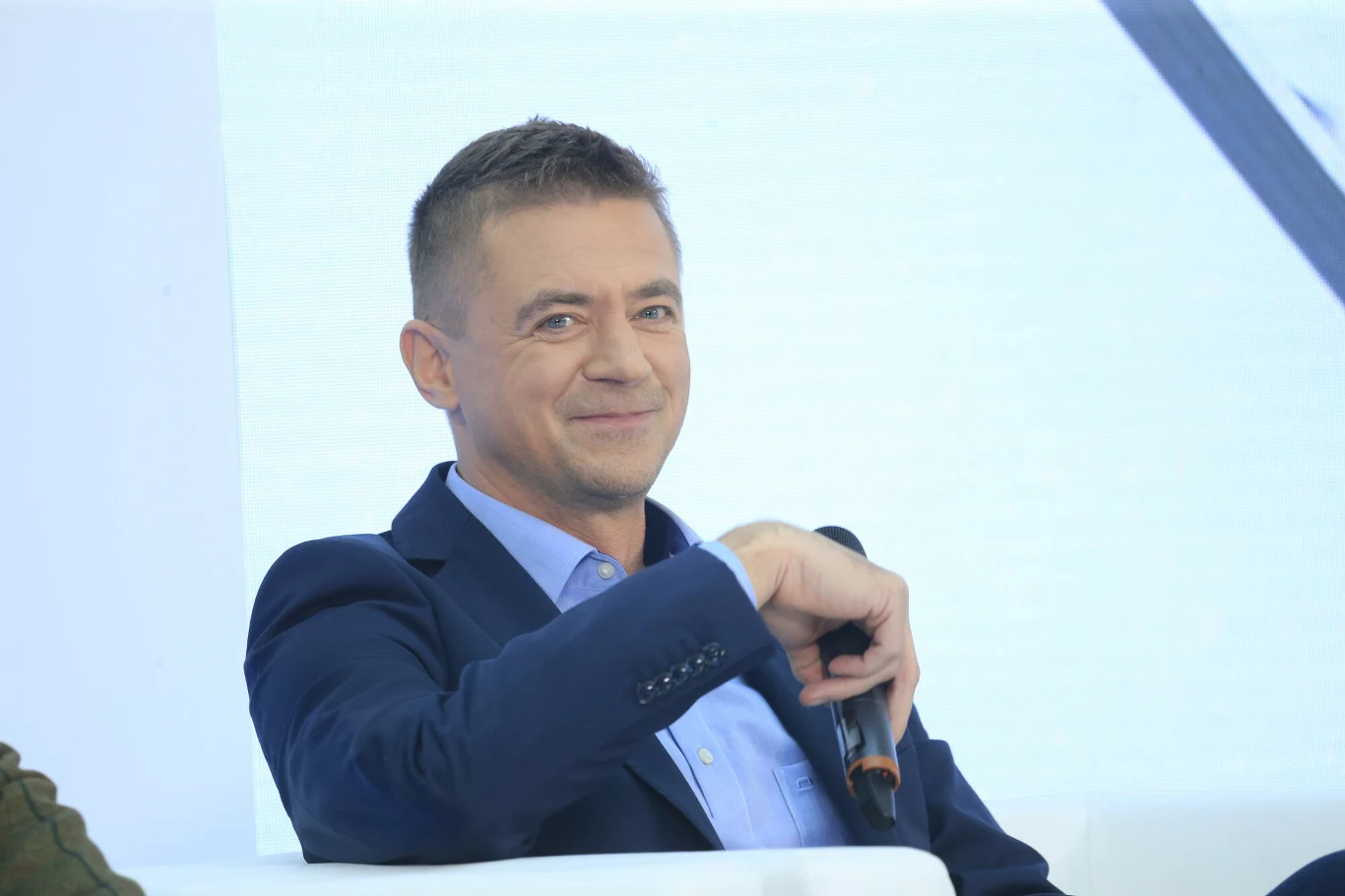 Igor Błachut Mężczyzna w garniturze siedzący na jasnej sofie, trzyma mikrofon i lekko uśmiecha się, patrząc w obiektyw aparatu. W tle jasne, minimalistyczne otoczenie.