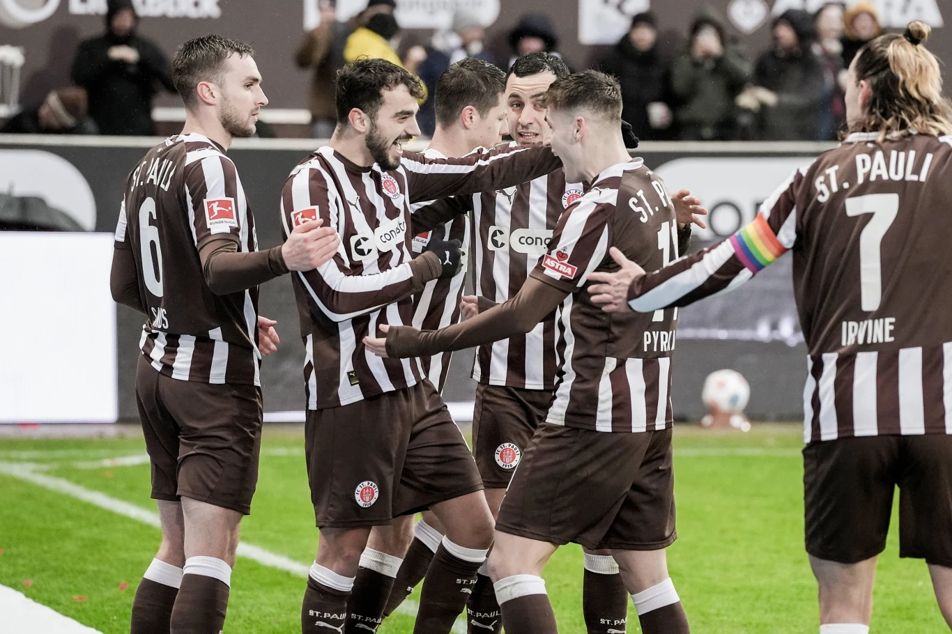Piłkarze drużyny St. Pauli w brązowo-białych strojach świętują zdobycie bramki na boisku, otoczeni przez kolegów z zespołu, jeden z zawodników nosi opaskę kapitana w kolorach tęczy.