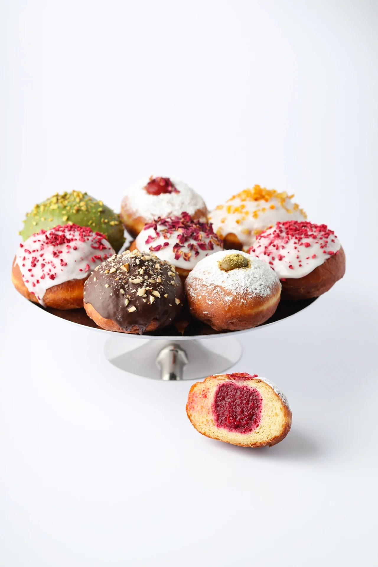 Różnorodne pączki z kolorowymi polewami i posypkami ułożone na metalowej paterze, jeden pączek przekrojony pokazuje nadzienie malinowe.