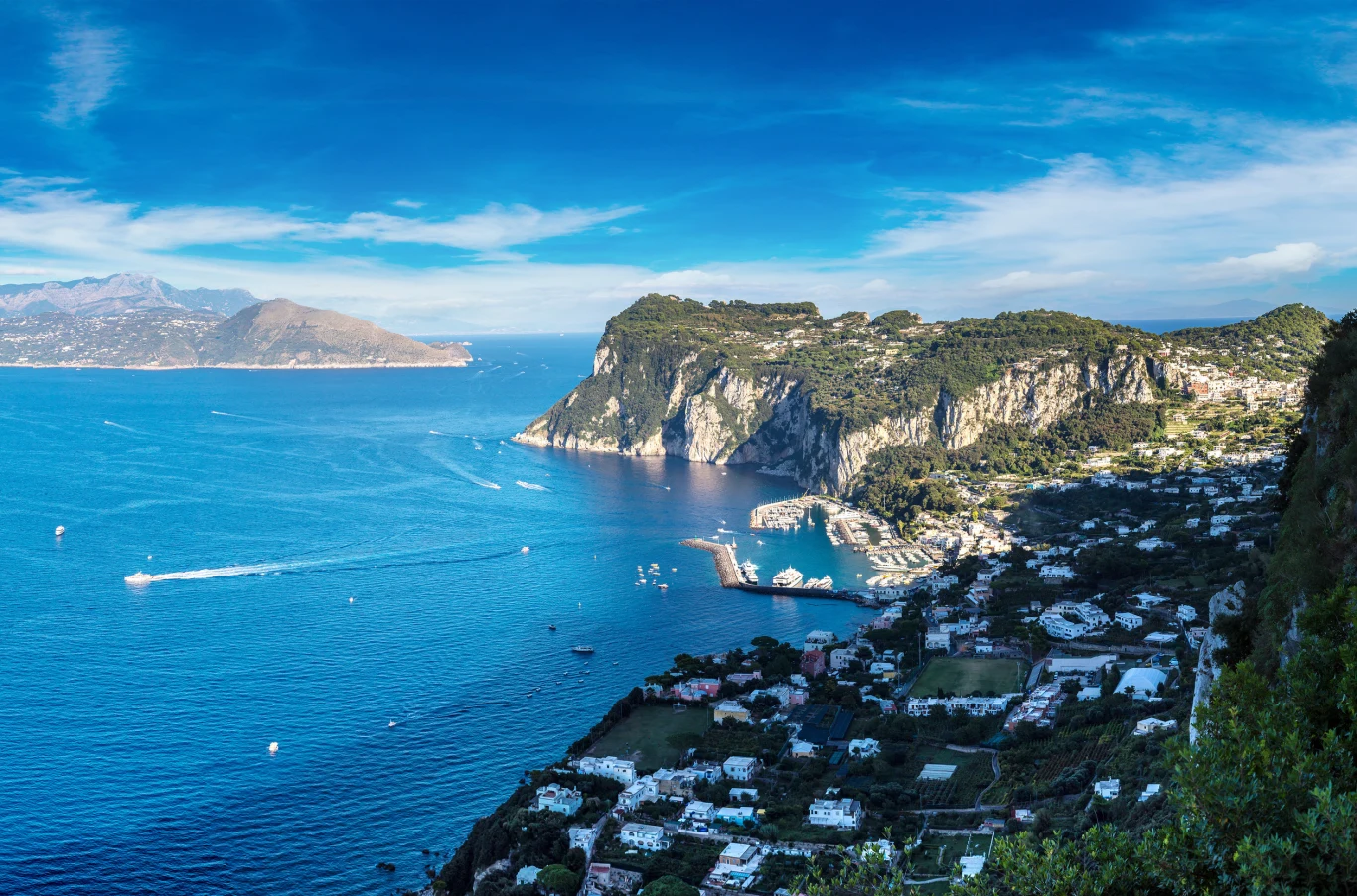 Rada włoskiej wyspy Capri wprowadza nowe ograniczenia dla turystów. Widok na lazurowe morze i malownicze klify otaczające zatokę z przystanią jachtową. Po prawej stronie zabudowania i zieleń na stokach, w tle wzgórza i wyspa przy wybrzeżu.
