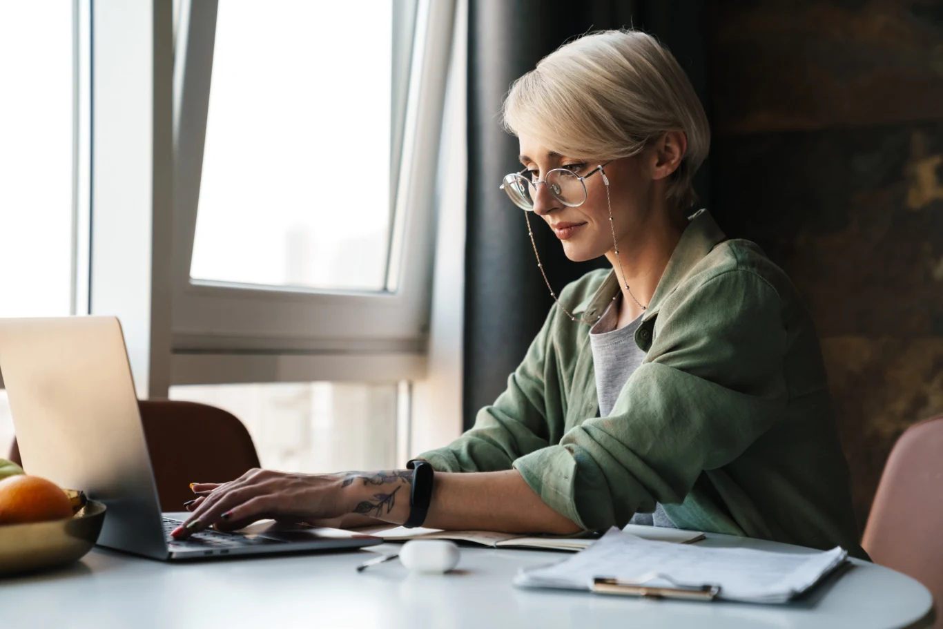 Kobieta z blond włosami i okularami pracuje przy komputerze przenośnym, skupiona na ekranie, siedzi przy stole, na którym leżą notatki, długopis i etui na słuchawki. Za nią znajduje się jasne okno z wpadającym światłem dziennym.