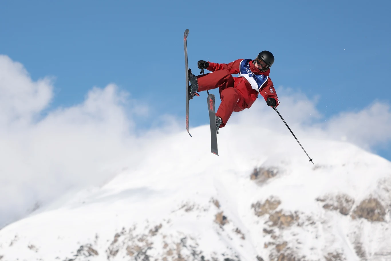 Narciarz freestyle wykonujący efektowny skok w powietrzu nad ośnieżonymi górami, ubrany w czerwony strój oraz kask ochronny, wyraźna dynamika oraz sportowy charakter ujęcia.