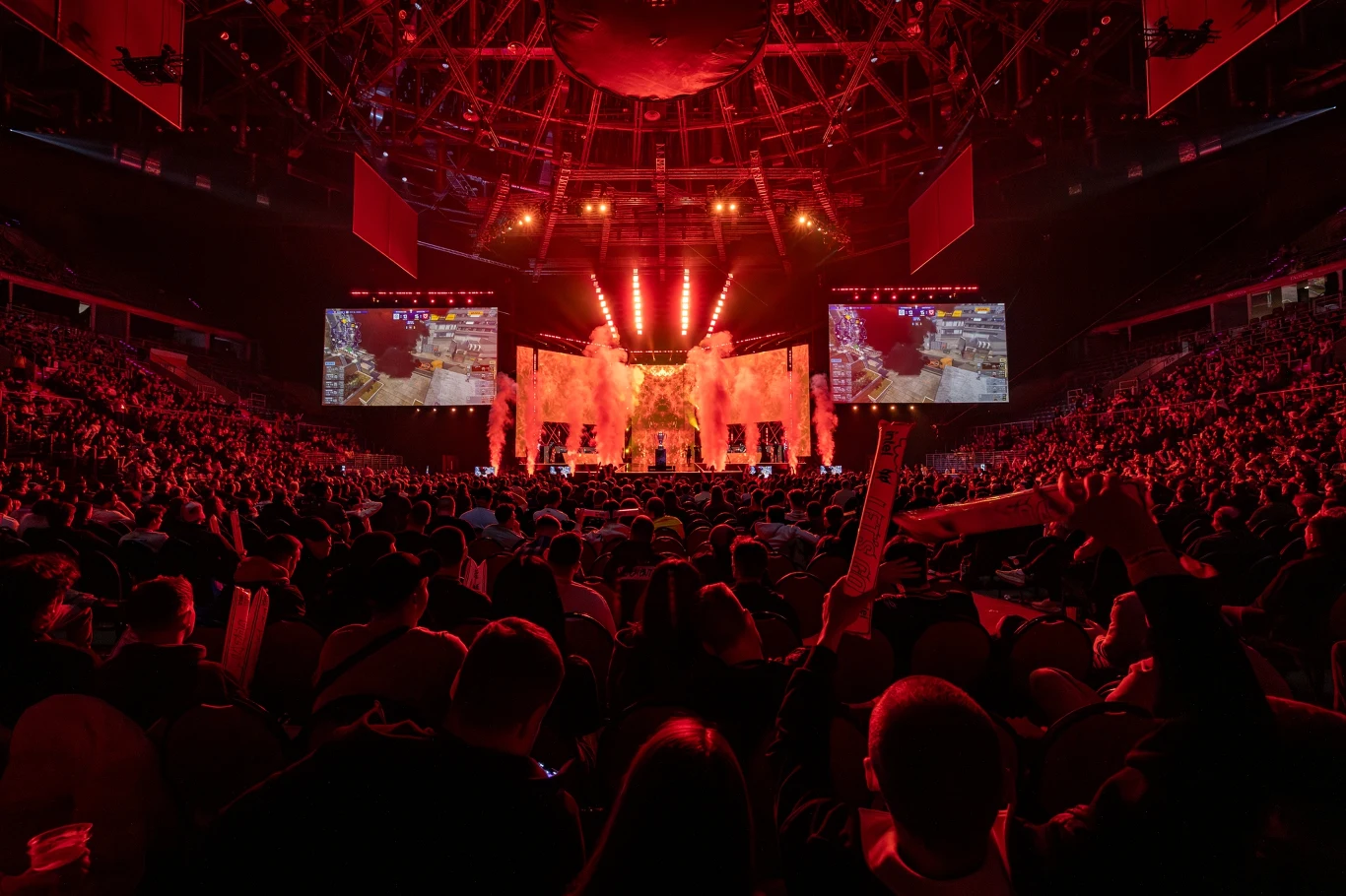 IEM 2026 Wielotysięczna publiczność w arenie kibicuje podczas emocjonującego wydarzenia e-sportowego, scena oświetlona czerwonymi światłami, duże ekrany transmitują rozgrywkę, na środku sceny widoczne efekty pirotechniczne.