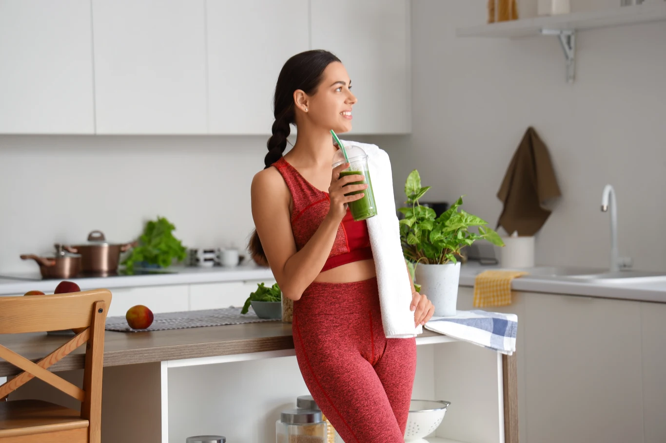 W tym roku skupisz się na sobie Młoda kobieta w czerwonym stroju sportowym opiera się o blat kuchenny, trzymając w dłoni zielony koktajl i ręcznik na ramieniu, wokół niej widoczne świeże warzywa i nowoczesne wnętrze kuchni.