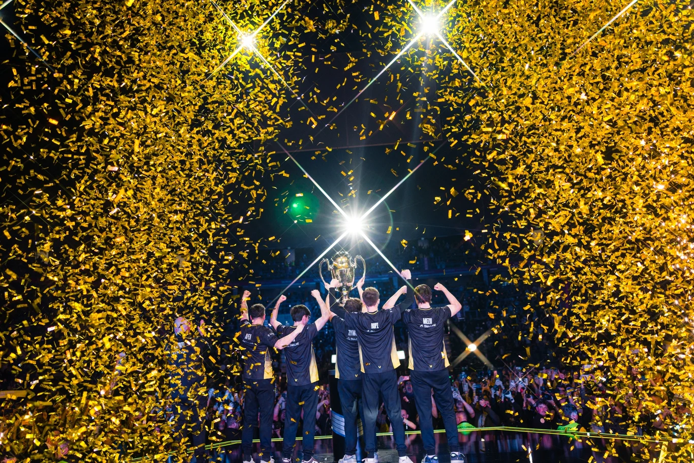 IEM 2026 Grupa młodych mężczyzn ubrana w jednakowe koszulki stoi na scenie i świętuje zwycięstwo, trzymając puchar w górze. W tle widoczny tłum kibiców oraz mnóstwo złotych konfetti spadających z góry, całość oświetlona jasnymi reflektorami.