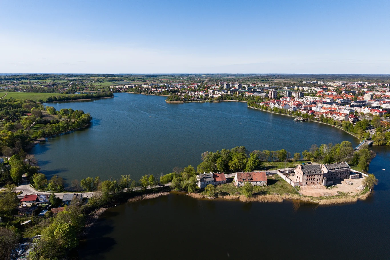 Bon turystyczny 2026 zachęca do odwiedzin malowniczego województwa warmińsko-mazurskiego Duże jezioro otoczone zielenią, w tle zabudowa miejska z charakterystycznymi kolorowymi dachami oraz fragmentem ruin czy zamku po prawej stronie na pierwszym planie.