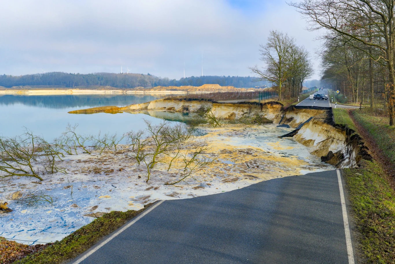 Droga częściowo zapadnięta i zalana wodą, zniszczona przez erozję brzegu jeziora, otoczona drzewami i rozlewiskami wodnymi, w tle linia lasu oraz chmury.