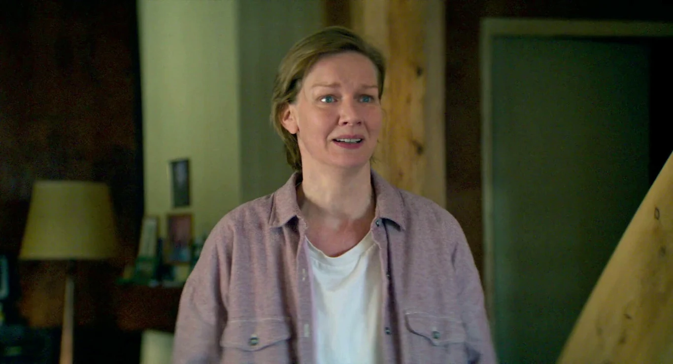 Sandra Hüller w filmie "Anatomia upadku" Kobieta w jasnej koszuli i białym t-shircie stoi we wnętrzu pokoju z drewnianą belką, w tle lampa oraz rozmazane elementy wyposażenia.