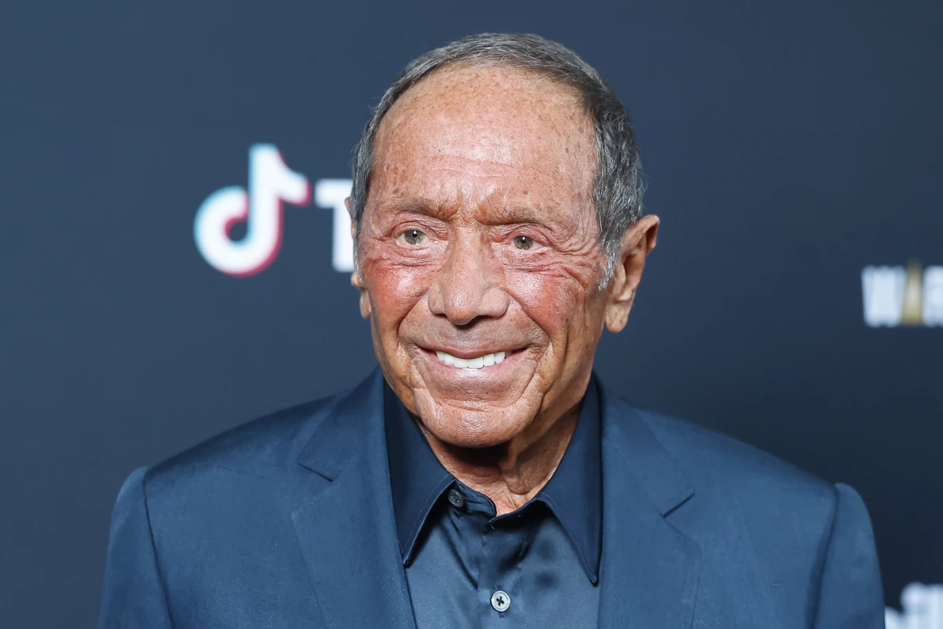 Paul Anka był trzykrotnie żonaty Starszy mężczyzna w ciemnym garniturze stoi na tle tablicy z logotypami, uśmiecha się delikatnie do kamery, w tle widoczny fragment logo TikTok.