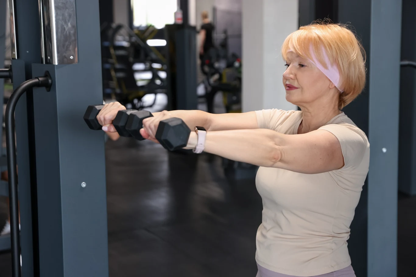 Starsza kobieta w jasnej koszulce i opasce na głowie ćwiczy z hantlami na siłowni; skupiona twarz świadczy o zaangażowaniu w wykonywane ćwiczenie, w tle widoczne są maszyny do ćwiczeń.