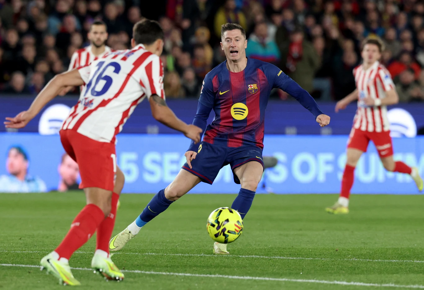 Dwóch piłkarzy podczas dynamicznej akcji meczu piłki nożnej, jeden w stroju FC Barcelona próbuje przejąć piłkę od zawodnika w biało-czerwonym stroju drużyny przeciwnej, w tle wyraźny stadion z publicznością.