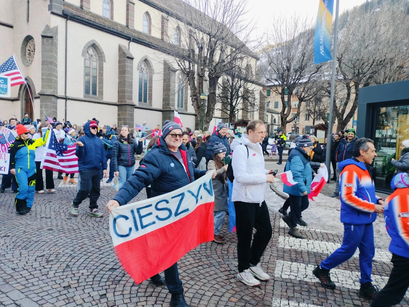 Tłum ludzi maszerujący ulicą w zimowych ubraniach, wiele osób trzyma flagi różnych krajów, w tym biało-czerwoną flagę z napisem 'CIESZYN'. W tle widoczny zabytkowy budynek kościoła oraz drzewa bez liści.
