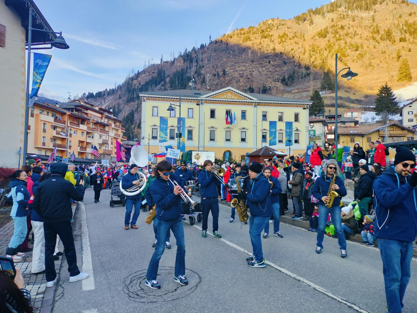 Orkiestra dęta maszerująca ulicą wśród licznie zgromadzonych widzów, kolorowe stroje uczestników oraz zimowe, górskie otoczenie, w tle zabytkowy budynek oraz stok porośnięty drzewami.