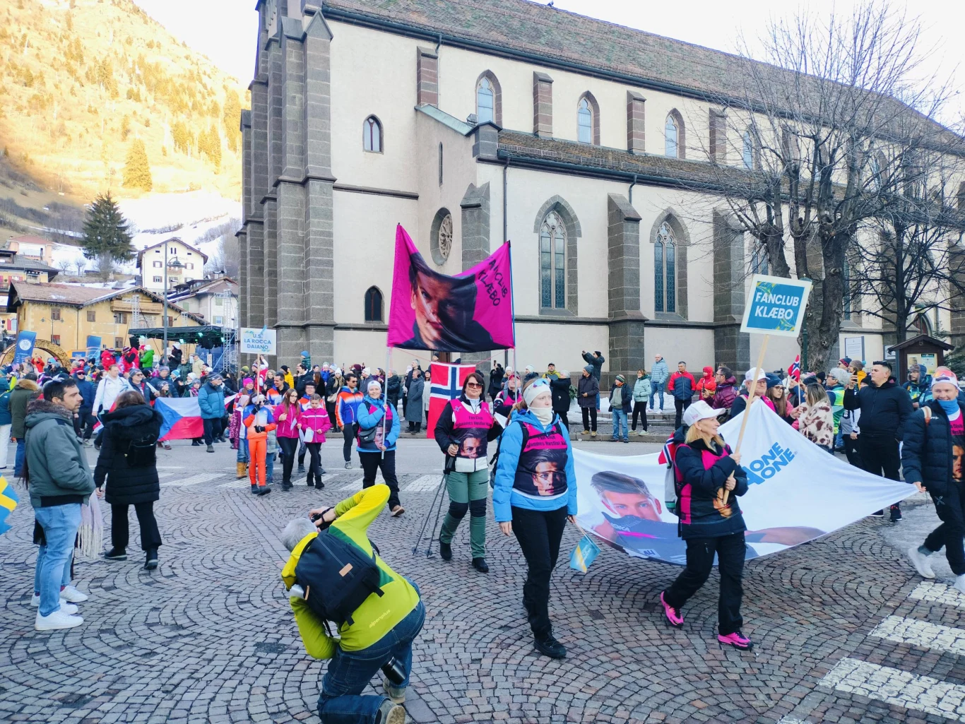 Grupa ludzi uczestnicząca w marszu lub manifestacji na brukowanym placu przed kościołem, trzymająca transparenty i flagi, otoczona przez osoby fotografujące i obserwujące. W tle widać historyczny budynek i drzewa bez liści, a uczestnicy ubrani są w zim...