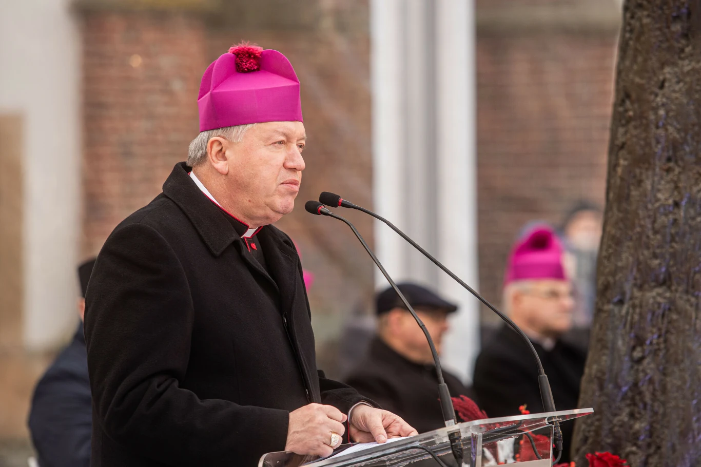 Archidiecezja wrocławska ostrzega przed oszustami podszywającymi się pod abp Józefa Kupnego Duchowny w stroju liturgicznym z fioletową piuską wygłasza przemówienie przy mównicy na otwartym powietrzu, w tle inni duchowni, również w fioletowych nakryciach głowy.