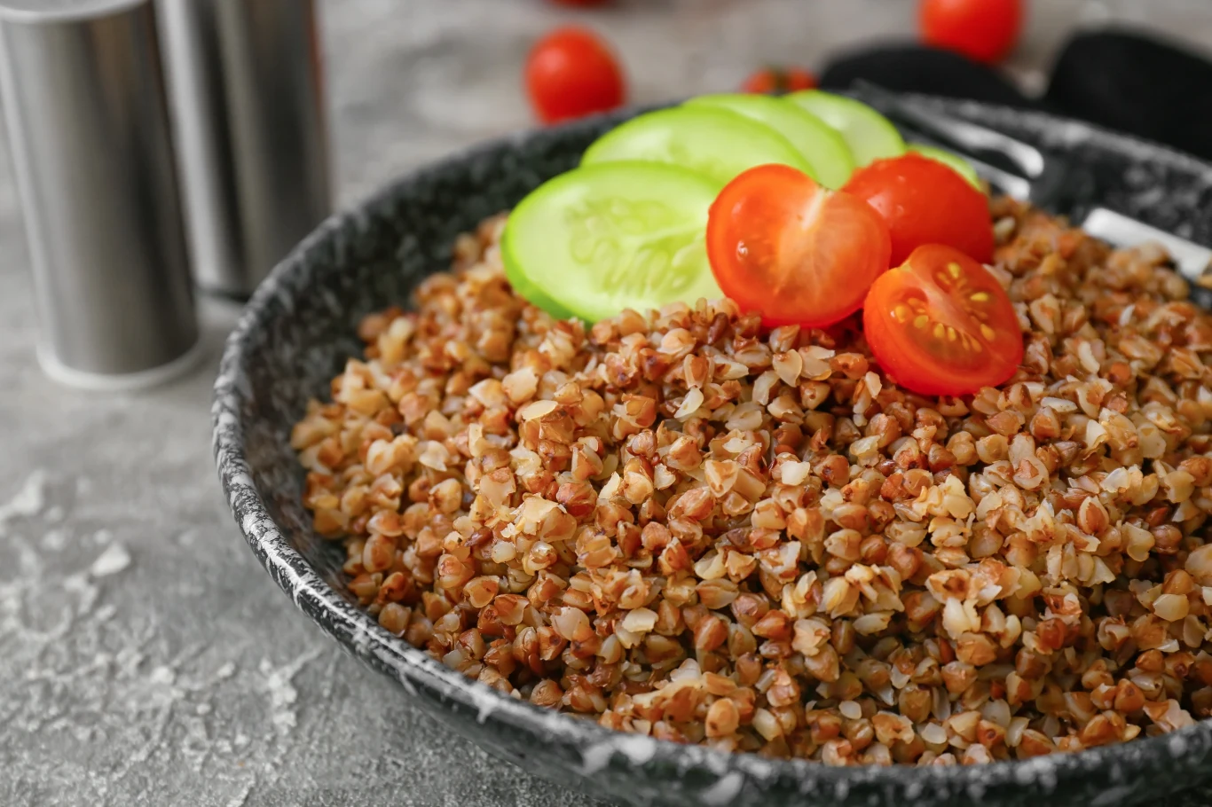 Kasza gryczana podana w głębokim talerzu, udekorowana świeżymi plasterkami ogórka i połówkami pomidorków koktajlowych.