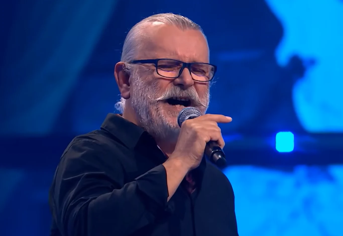 Artur Słowik w "The Voice Senior" Starszy mężczyzna z siwą brodą i wąsami, w okularach i czarnej koszuli, śpiewa do mikrofonu na tle sceny oświetlonej na niebiesko.
