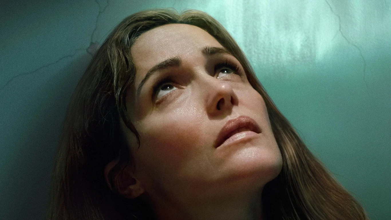 Rose Byrne w filmie "Kopnęłabym cię, gdybym mogła" Twarz kobiety skierowana ku górze, wyraz twarzy sugeruje napięcie lub zamyślenie, jasne światło pada na czoło, w tle subtelnie widoczna ściana z pęknięciem.