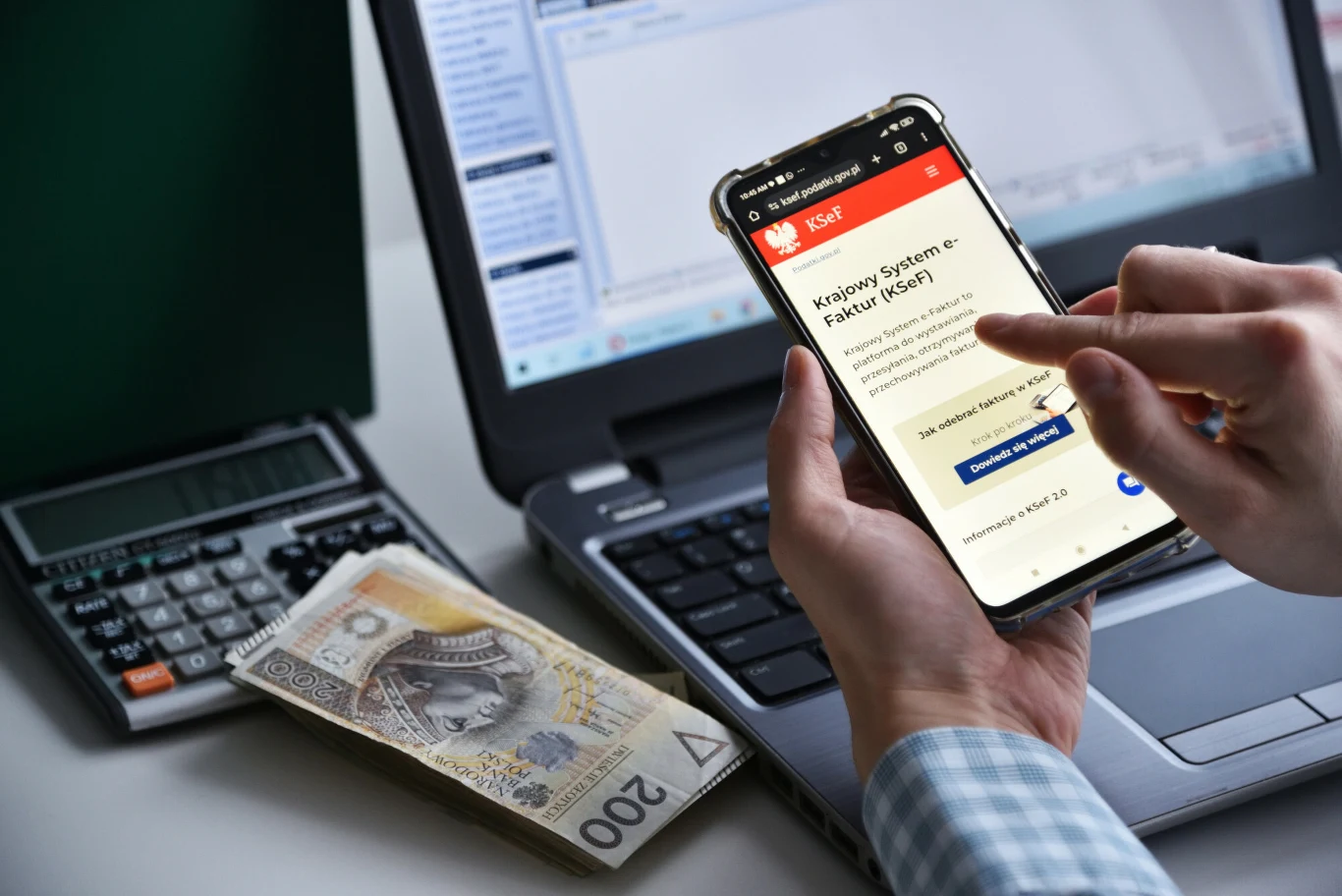 KSeF stał się obowiązkowy dla części firm od lutego Ręka trzymająca smartfona z otwartą stroną Krajowego Systemu e-Faktur na tle laptopa, obok leży kalkulator i polski banknot dwustuzłotowy.