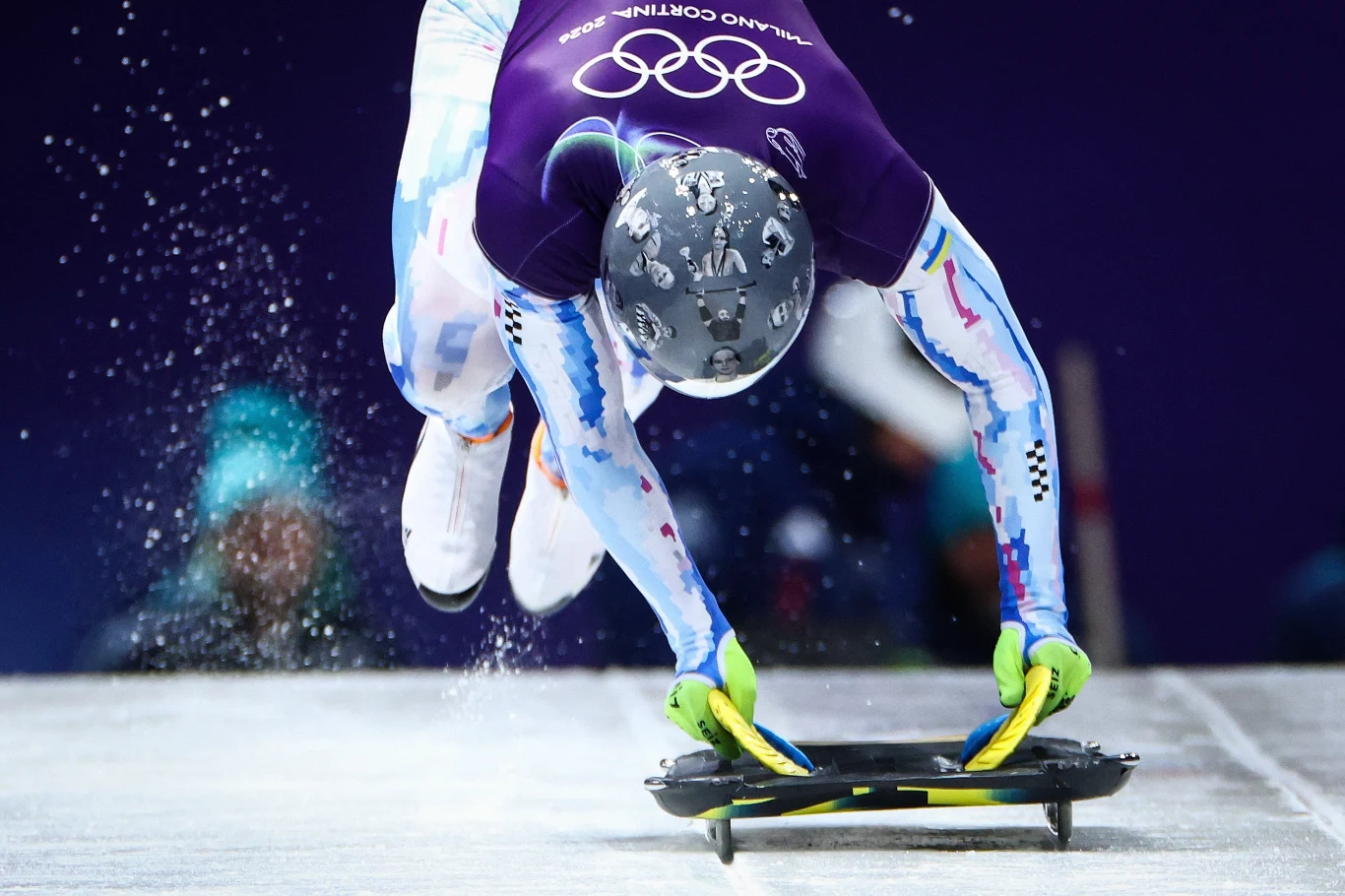 Władysław Heraskiewicz Sportowiec ubrany w strój ochronny podczas startu w konkurencji skeletonu na lodowym torze, w tle logo igrzysk olimpijskich.